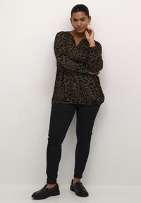 Leopardmuster Langarmshirt in Braun und Schwarz, kombiniert mit schwarzen Skinny Jeans. Das Model trägt glänzende schwarze Schuhe und zeigt eine lässige Passform.