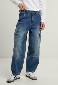 Baggy blue denim cargo pants with multiple pockets and a faded finish. Fabric is sturdy with a smooth texture, featuring a button and zip closure.