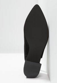 Semelle de chaussure noire à bout pointu reposant verticalement contre un mur d’angle blanc, montrant un talon texturé et une zone antérieure lisse.