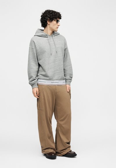 Uomo con occhiali da sole, felpa grigia con cappuccio, pantaloni beige a gamba larga e scarpe nere, in piedi e che guarda di lato.