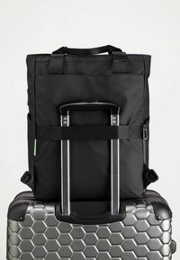 Svart tygryggsäck med topphandtag, utrustad med en resväska-rem. Lays på en silverresväska med hexagonmönster.