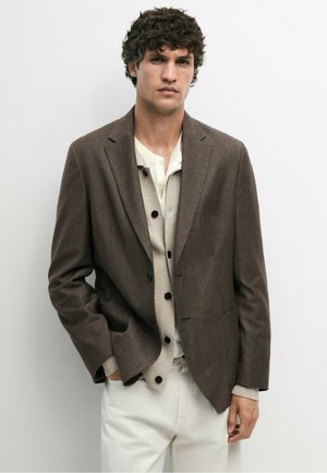 Man met krullend donker haar, die een bruine blazer, beige gebuttoncardigan, wit overhemd en witte broek draagt tegen een effen achtergrond.