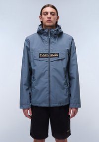 Chaqueta impermeable azul con cremallera frontal, capucha ajustable y un parche de marca en el pecho. Combinada con pantalones cortos negros.