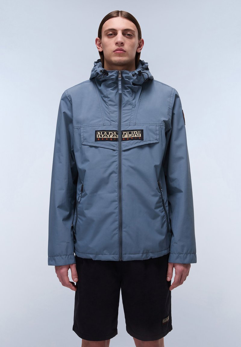 Chaqueta impermeable azul con cremallera frontal, capucha ajustable y un parche de marca en el pecho. Combinada con pantalones cortos negros.
