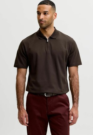 Mand iført en mørkebrun kortærmet poloshirt med lynlåslukning ved kraven og mørkerøde bukser mod en ensfarvet baggrund.