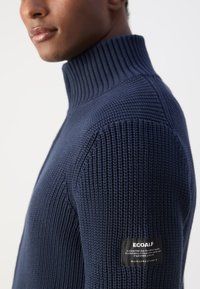 Maglione a collo alto lavorato a maglia di colore blu navy con texture a coste, dotato di un'etichetta nera ECOALF sulla manica e una silhouette aderente.