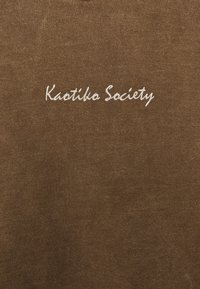 Smeđi krojački košulja s bijelim vezom na kojem piše "Kaotiko Society." Materijal izgleda mekano i teksturirano, bez uzoraka.