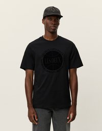 Čierne bavlnené tričko s okrúhlym logom v strede, ktoré obsahuje text "LES DEUX COPENHAGEN." Model má na hlave čiernu čiapku s textúrovaným vzorom.