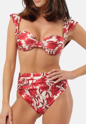 Donna che indossa un bikini a vita alta con stampa floreale di rose rosse e dettagli arricciati sulla parte superiore e inferiore su sfondo neutro.
