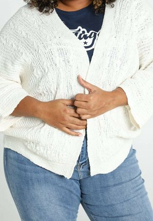 Personne portant un cardigan blanc en maille par-dessus une chemise bleu marine et un jean bleu, fermant le devant du cardigan avec les deux mains.