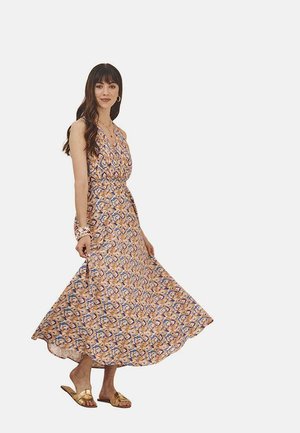 Vestido maxi de tela estampada con remolinos en naranja, azul y crema. Tiene un diseño de cuello halter y un movimiento de falda acampanada. Sandalias doradas.