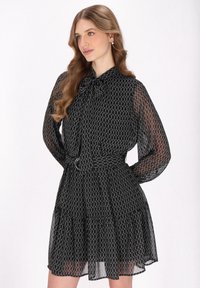 Zwart gestippeld jurk van doorzichtig materiaal, met een striknek, lange mouwen en een gelaagde rok met een ceintuur op de taille.