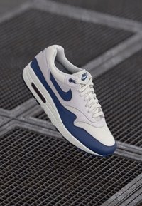 Nike Air Max 1 tenisice imaju bijeli i mornarsko plavi dizajn s teksturiranim gornjim dijelom, kopčanjem na vezanje i vidljivim zračnim jastučićima u potplatu.