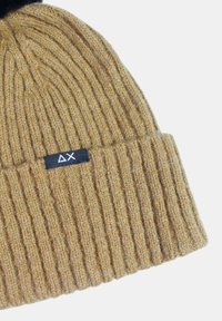 Beanie color beige a coste con risvolto e piccolo etichetta nera in tessuto con simboli triangolo e X bianchi.