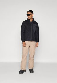 Črno jopico iz flisa z zadrgo in žepom na prsih; kombinirano z bež cargo hlačami in črnimi škornji. Moški z očali za sonce, stoji pokonci.