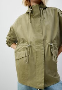 Jachetă verde oliv cu fermoar în față, șnururi ajustabile și două buzunare frontale. Fabricată dintr-un material ușor, neted.