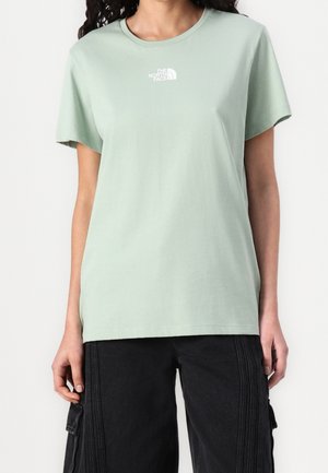 Woman wearing a light green The North Face T-shirt and black cargo pants, standing against a plain white background.