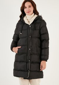 Black puffer coat featuring a hood, contrasting cream lining, button closures, and elastic cuffs, with visible quilted texture and side pockets.