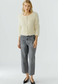 Cremfarbener Fake-Fur-Cardigan mit rundem Ausschnitt, langen Ärmeln und vorderem Knopfverschluss. Getragen mit grauen High-Waist-Jeans und schwarzen Ballerinas.