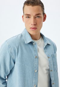 Lichtblauwe spijkerjacket met een knoopsluiting aan de voorkant, spitse kraag en één borstzak. Ggedragen over een witte T-shirt, zachte textuur.