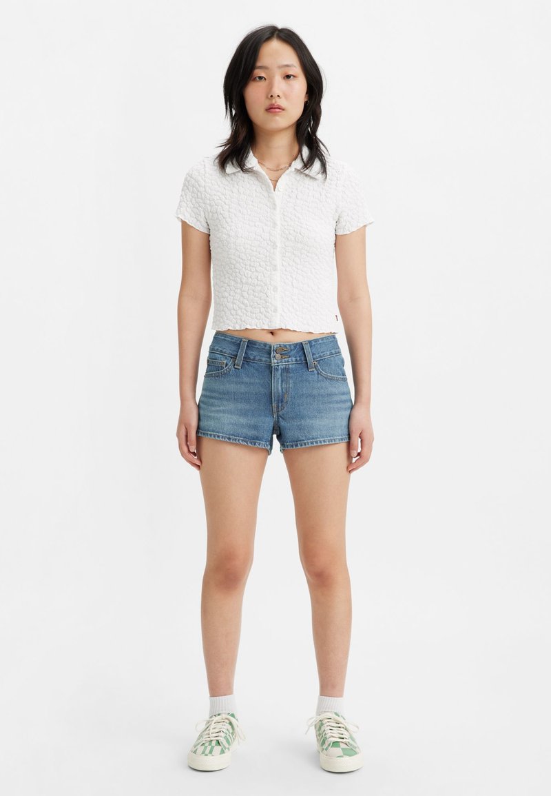 Chemise blanche texturée à manches courtes et coupe courte, assortie à un short en denim bleu et des baskets rayées vertes. Fond neutre.