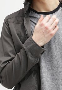 Person klädd i en grå jacka över en grå skjorta, med höger hand lyft och rör vid kragen.