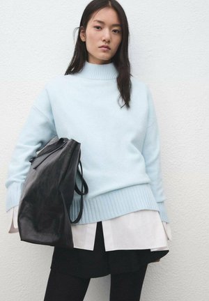 Pull bleu clair à col montant, ourlet côtelé et épaules tombantes, associé à un sac à dos en cuir noir et une chemise blanche à plusieurs couches.