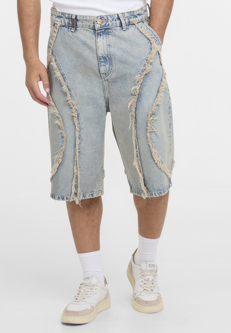 Homme portant un short en jean délavé clair au niveau des genoux avec des détails en patch effilochés, des chaussettes blanches et des baskets blanches sur un fond uni.