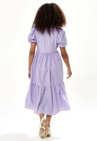 Lavender puff-sleeve dress with a tiered, ruffled skirt and elastic waist. Model wears white sneakers, showing a relaxed and casual fit.