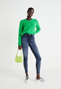 Vihreä pörröinen neule, siniset kapeat farkut, harmaat lenkkarit ja pieni limevihreä käsilaukku. Teksturoitu neulosta, jossa on V-pääntie ja pitkät hihat.