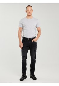 Mand stående iført en lysegrå kortærmet t-shirt, sorte revnede jeans med bælte og sorte snørestøvler på en ensfarvet baggrund.