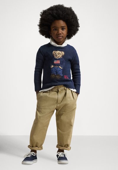 Mørk blå strikkegenser med et design av en teddybjørn med et amerikansk flagg, kombinert med khakifargede bukser og marineblå joggesko.