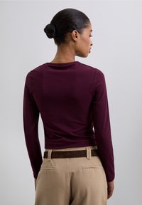 Long-sleeve fitted top in deep burgundy with a round neckline. Paired with light beige pants featuring a brown suede belt.