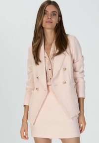 Helle rosa doppelt geknöpfte Blazer mit fransigen Kanten, goldenen Knöpfen und strukturiertem Stoff. Getragen über einer Spitzentunika und einem passenden Rock.