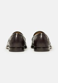 Un par de mocasines de cuero trenzado de color marrón oscuro visto desde atrás sobre un fondo blanco liso.