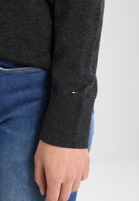 Gros plan sur une main à côté du poignet d'un pull à manches longues gris foncé avec un petit logo brodé rouge, blanc et bleu, associé à un jean bleu.