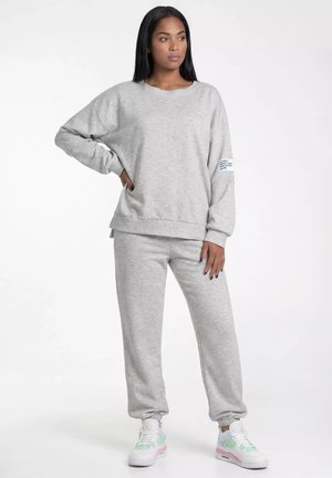 Kobieta stojąca z jedną ręką na biodrze, ubrana w jasnoszarą bluzę i dopasowane spodnie dresowe z białymi trampkami na białym tle.