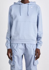Sweat à capuche bleu clair avec cordons associé à un pantalon cargo assorti avec poches latérales, porté par une personne debout sur un fond uni.
