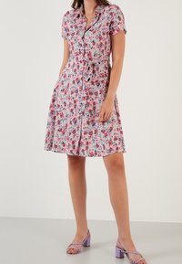 Květinové košilové šaty v růžové barvě s páskem v oblasti pasu. Krátké rukávy, délka po kolena, vyrobené z lehkého materiálu s vzorem růží a listů.