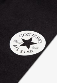 Μαύρο ύφασμα με λευκό κυκλικό λογότυπο. Το λογότυπο περιλαμβάνει ένα μαύρο αστέρι και κείμενο: "CONVERSE", "All Star" και "Chuck Taylor".