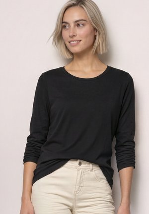 Lyshåret kvinde med kort hår iført en sort langærmet skjorte og lyse beige bukser, stående mod en neutral baggrund.