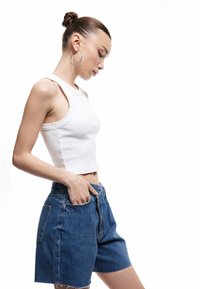 Nuori nainen, jolla on nuttura, päällä valkoinen olkaimeton crop-top, siniset farkkushortsit, suuret korvakorut ja toinen käsi taskussa.