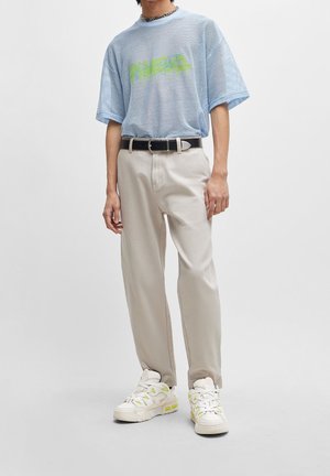 Light blue mesh t-shirt with green graphic, beige trousers, black belt, and cream sneakers with green accents. Casual fit and sporty design.