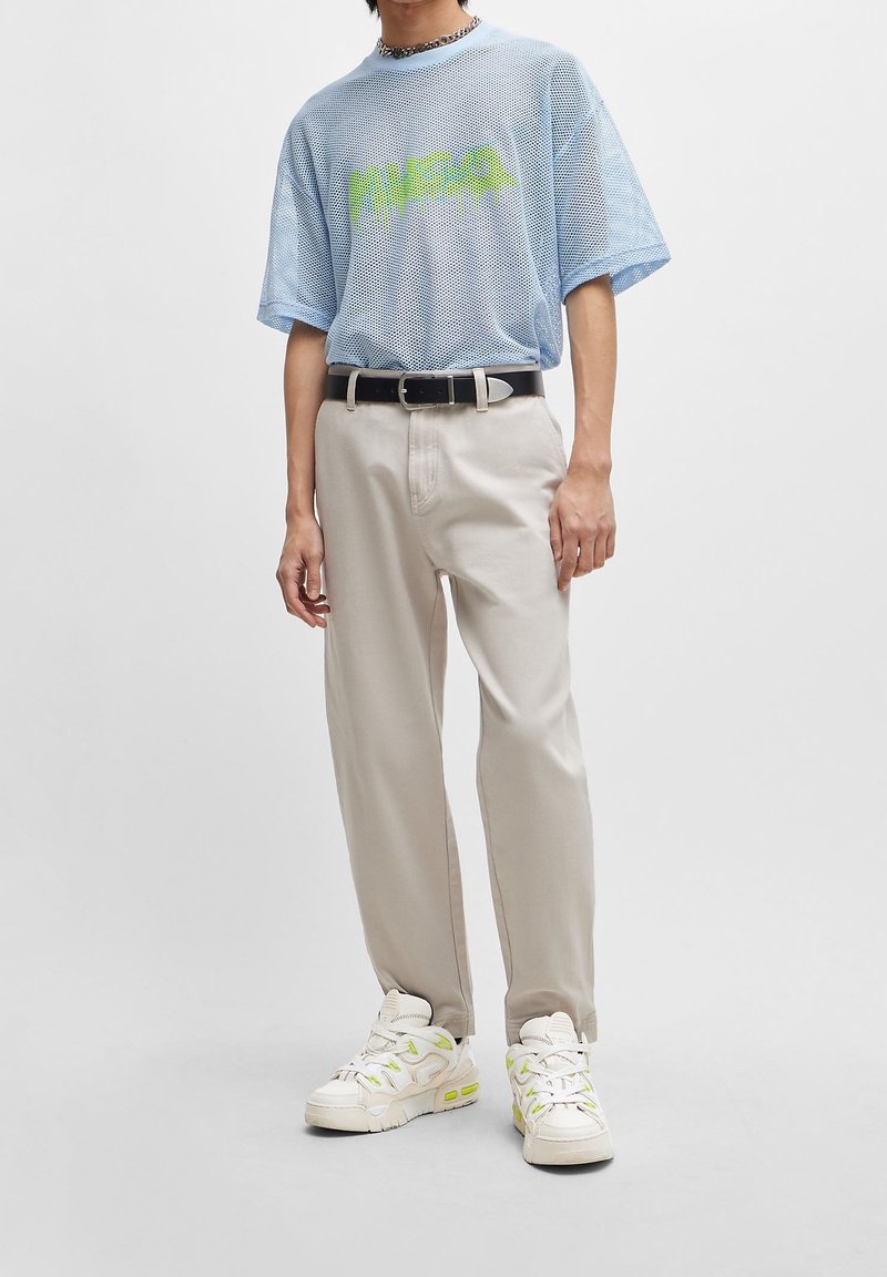 T-shirt in rete blu chiaro con grafica verde, pantaloni beige, cintura nera e scarpe da ginnastica crema con dettagli verdi. Fit casual e design sportivo.