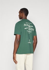 Πράσινο βαμβακερό μπλούζα με κοντό μανίκι, με μεγάλη γραφική εκτύπωση στην πλάτη, που περιλαμβάνει κείμενο και εικονογραφήσεις.