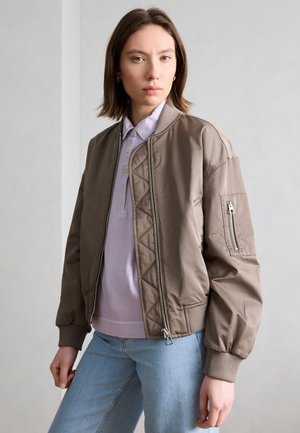 Mladá žena s krátkými hnědými vlasy, která má na sobě taupe bomber bundu přes lila polo tričko a světle modré džíny, stojící před jednoduchou zdí.