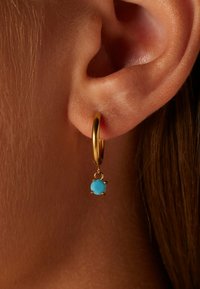 Gouden oorbel met een blauwe edelsteen pendant. De steen is rond en gezet in een decoratieve setting, hangend onder de ring.