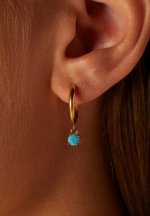 Gouden oorbel met een blauwe edelsteen pendant. De steen is rond en gezet in een decoratieve setting, hangend onder de ring.