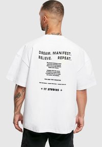 Biely oversized tričko s čiernym textom na zadnej strane, obsahujúce motivačné frázy a značku "2Y STUDIOS"; bavlnený materiál, voľný strih.
