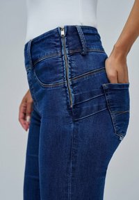 Vaqueros de mezclilla azul oscuro con cremallera lateral, diseño ajustado y un bolsillo de parche; la tela tiene una textura suave con sutiles acentos de costura.